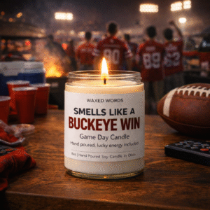 Waxed Words “Smells Like a Buckeye Win” candle on a wooden table with football, red cups, and fans under stadium lights at a nighttime tailgate.