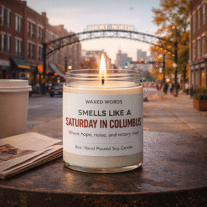 Waxed Words “Smells Like a Saturday in Columbus” candle on a café table with coffee and newspaper, Short North arches in the background.