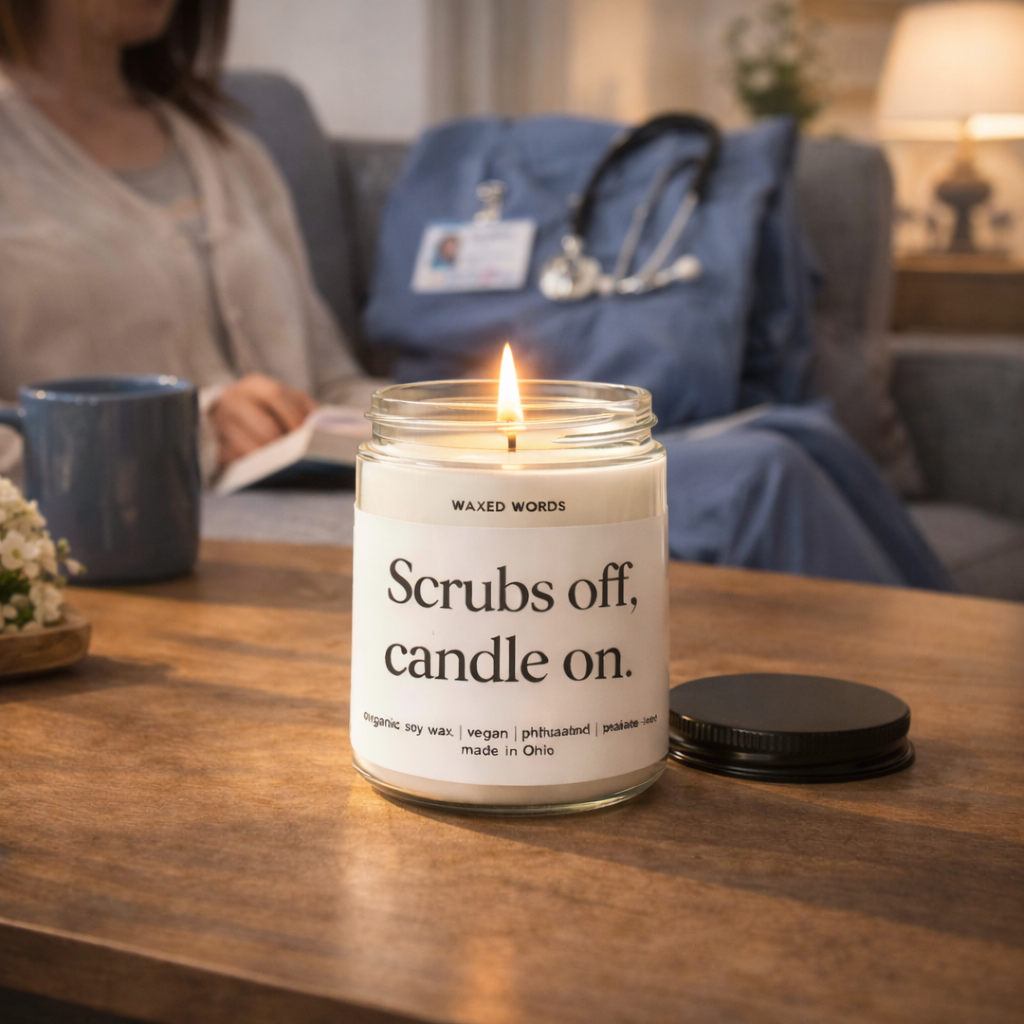 Waxed Words soy candle with “Scrubs off, candle on” label on a wooden table in a nurse’s living room with scrubs and stethoscope on the couch in the background.